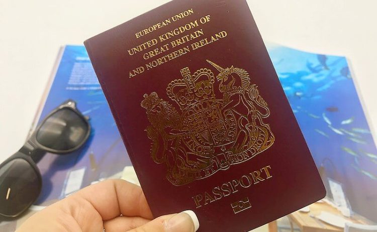 A red UK European passport in focus, in front of a travel brochure and some black sunglasses. Photo by Holiday Gems. Flickr.
